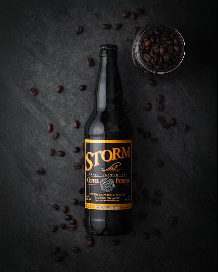 Dark artisanal coffee beer bottle surrounded by coffee beans on a dark surface.